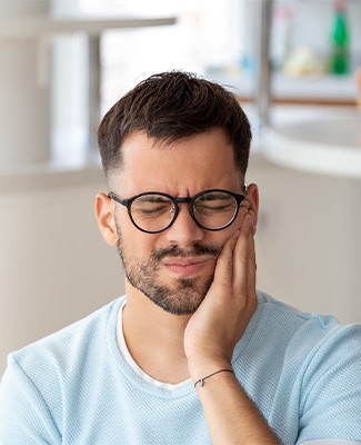 Man in black-rimmed glasses holding hand to face in pain